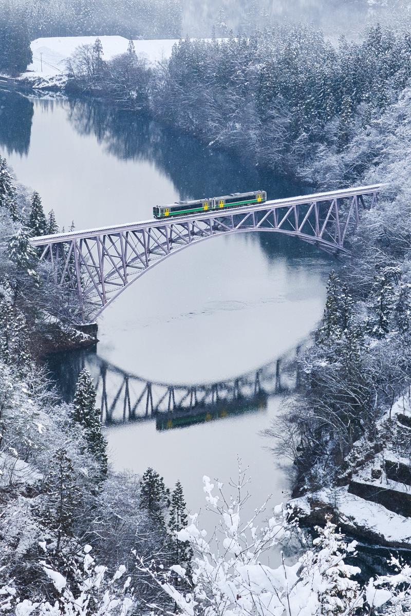 A train with green and yellow stripes running on a snow-covered railway bridge crossing over a snowy valley
