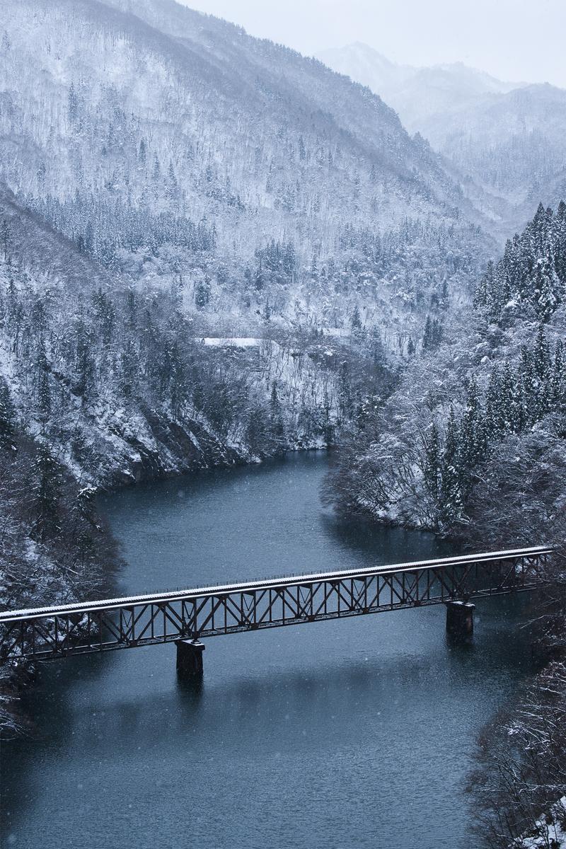 A reddish-brown steel-frame bridge crossing over a river, with snow-covered and tree-covered mountains on both sides