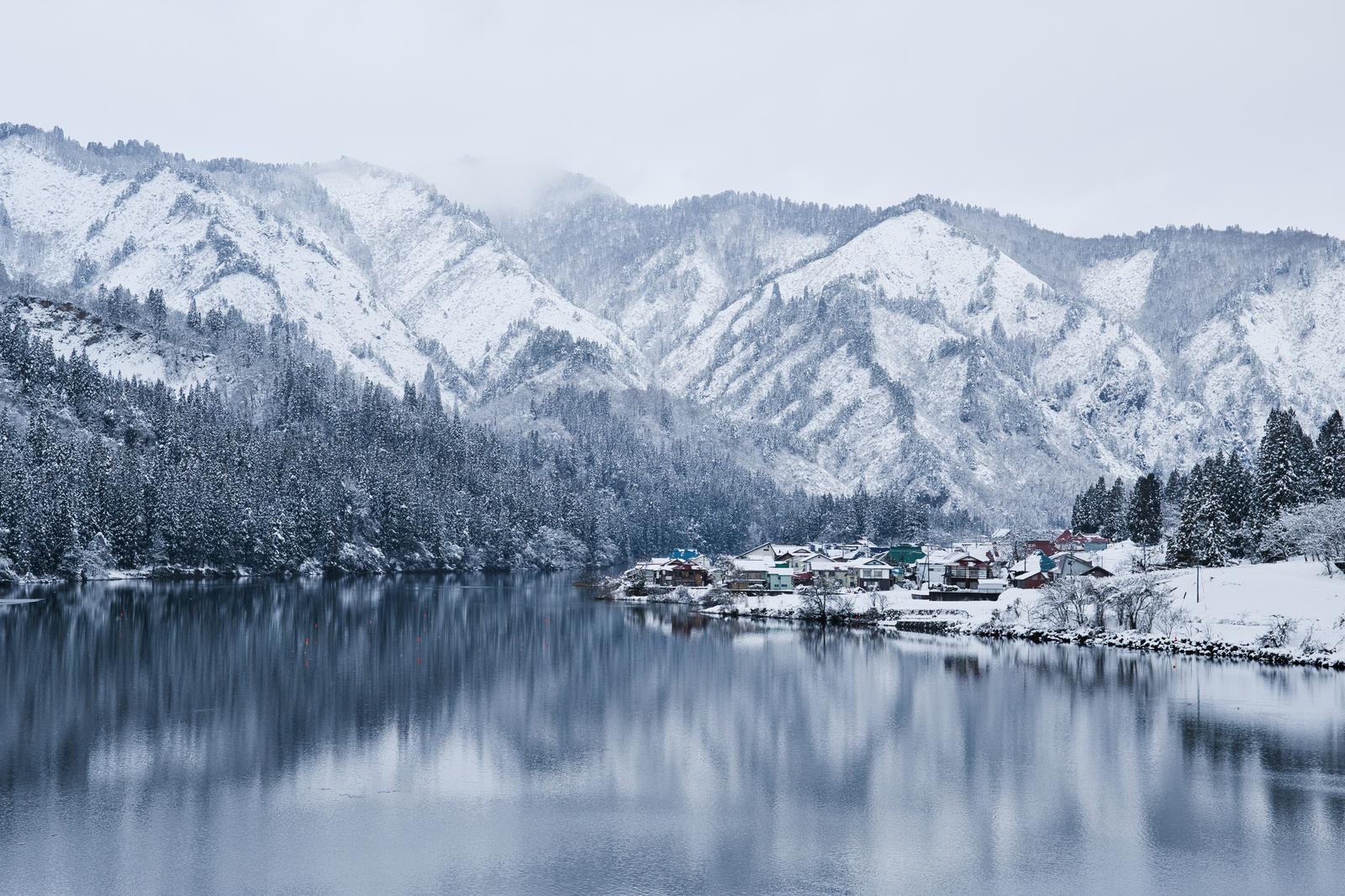 A winter landscape of the snow-covered Odaishimura settlement along the Tadami River, with snow-capped mountains in the background.