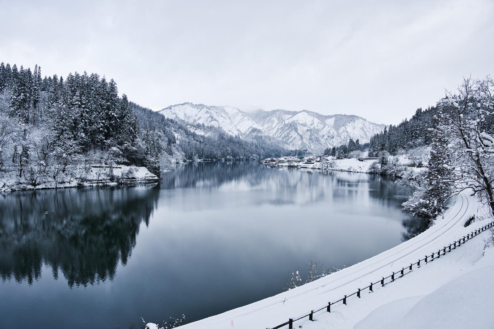 A river that reflects the snow-covered coniferous forest and distant snow-capped mountains. A white and black fence runs through the snow in the foreground.