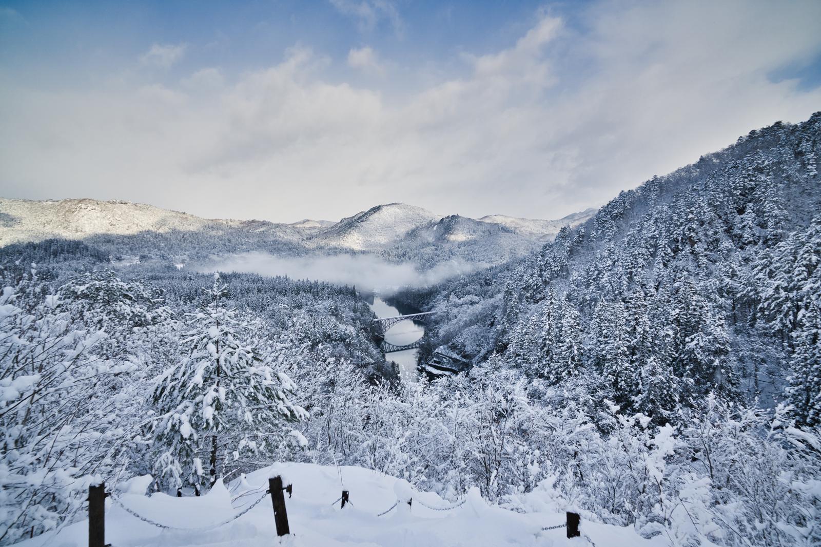 A white bridge spans the valley surrounded by snow-covered, coniferous tree-covered mountains in the winter