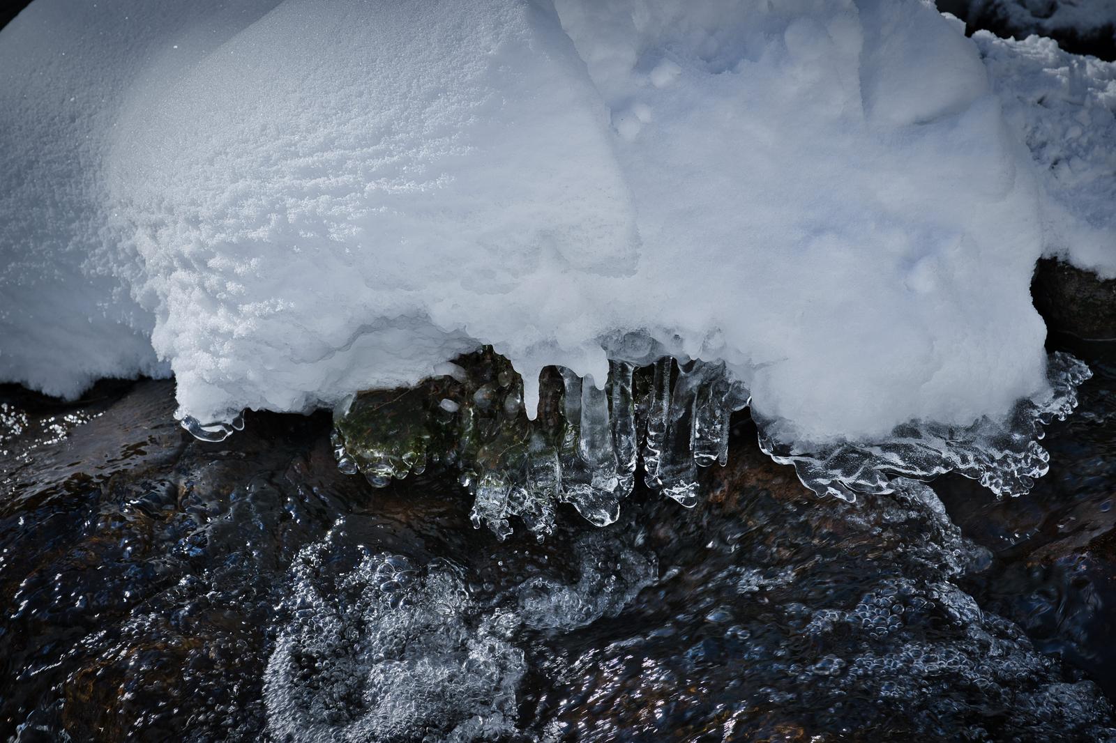 Close-up of translucent icicles hanging from a thick snow overhang above dark wet rocks, with fast-moving stream water flowing beneath in winter.