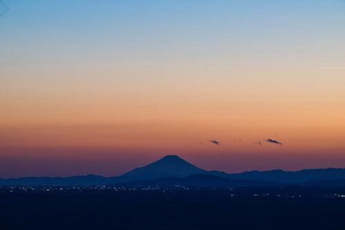 オレンジ色の夕焼けに浮かぶ富士山のシルエット、手前に町の灯りの写真