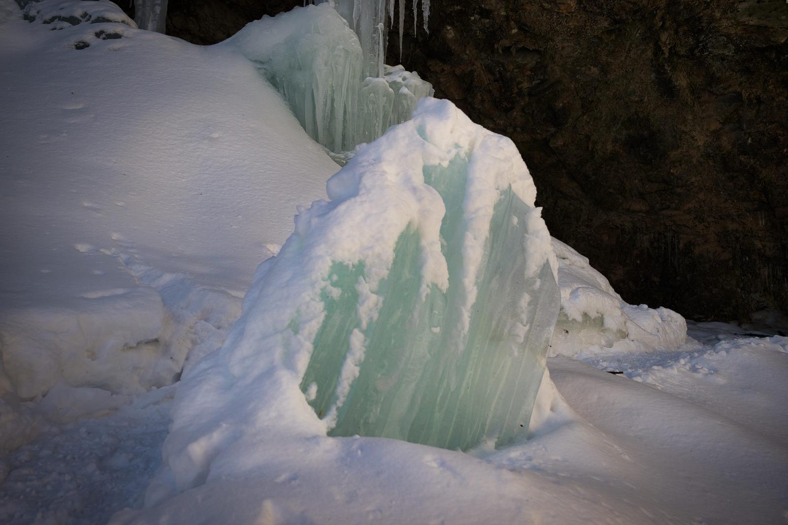 The winter scenery with thick, white snow layered and overlapping with thin, greenish-colored ice, and icicles hanging down.