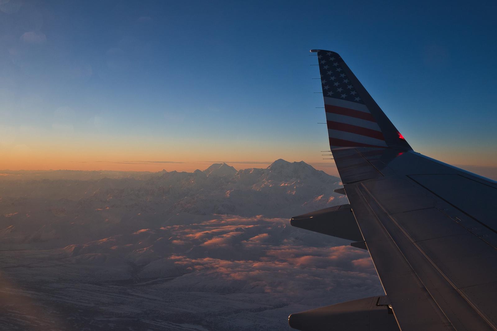 飛行機の窓から見える米国旗の翼と、夕焼けに染まった雪山と川の風景