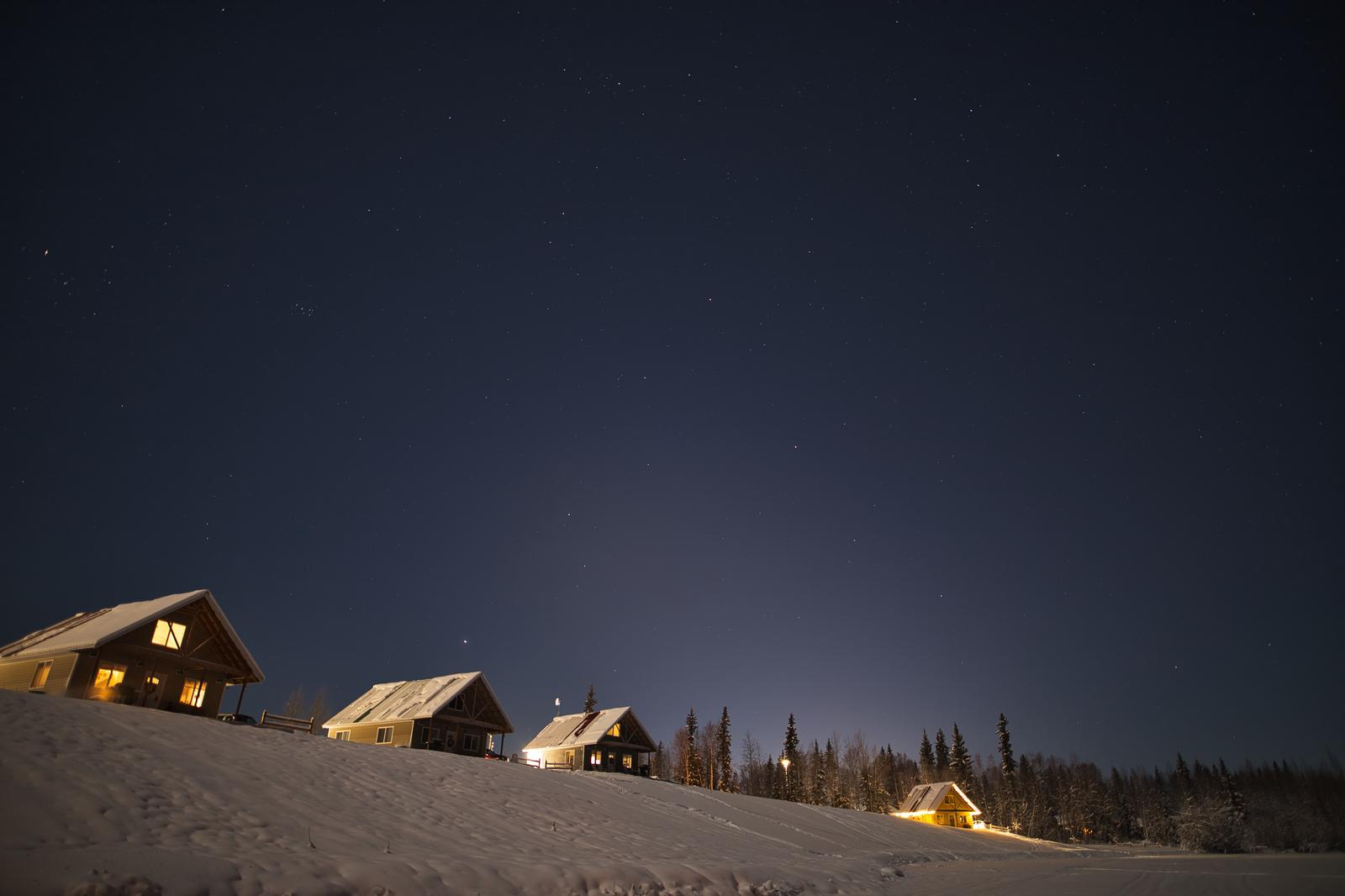 雪に覆われた丘の上に灯りのついた複数の山小屋が建ち、星空が広がる夜景