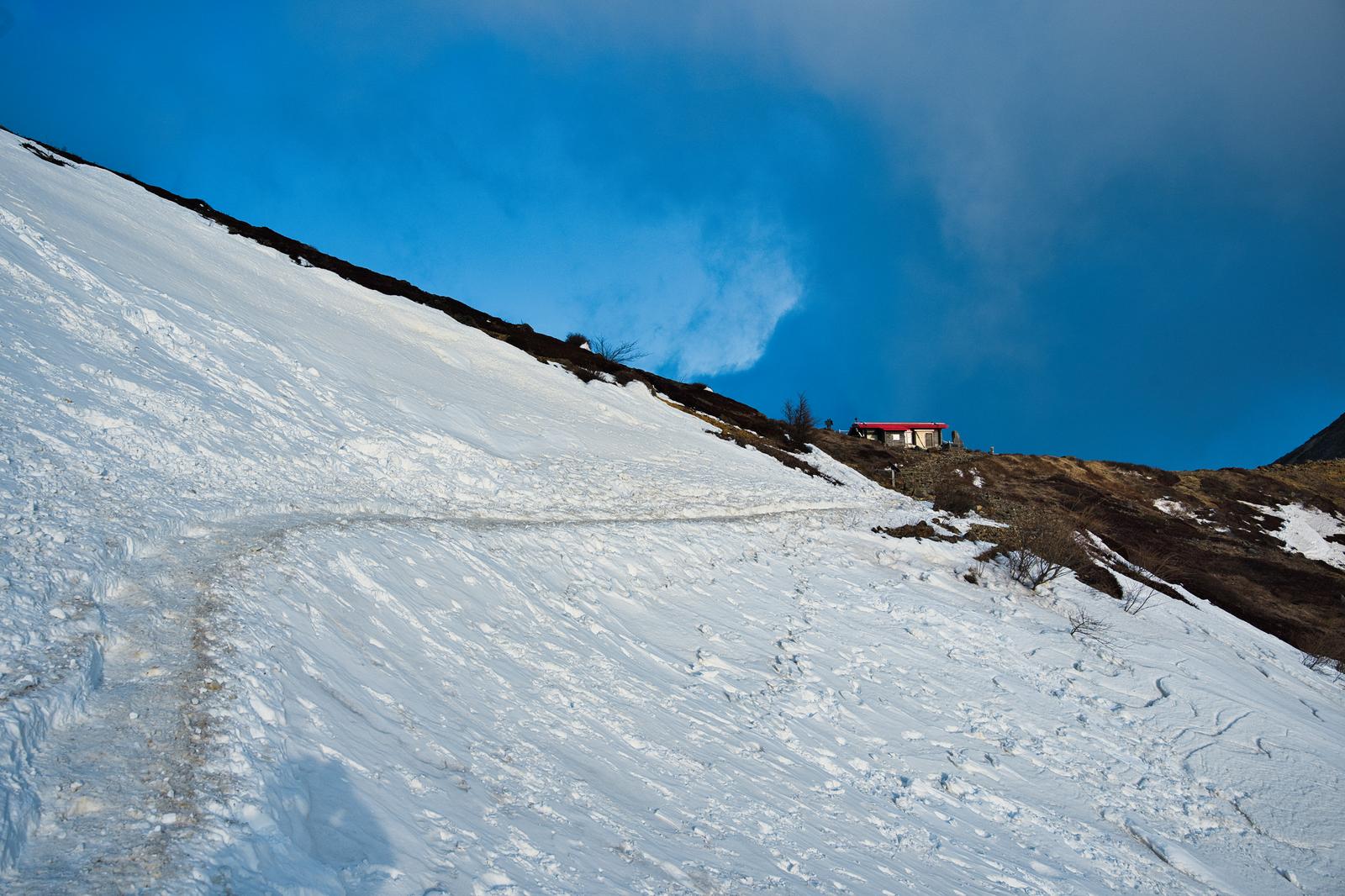残雪の斜面に続く登山道の先、稜線に建つ赤い屋根の山小屋