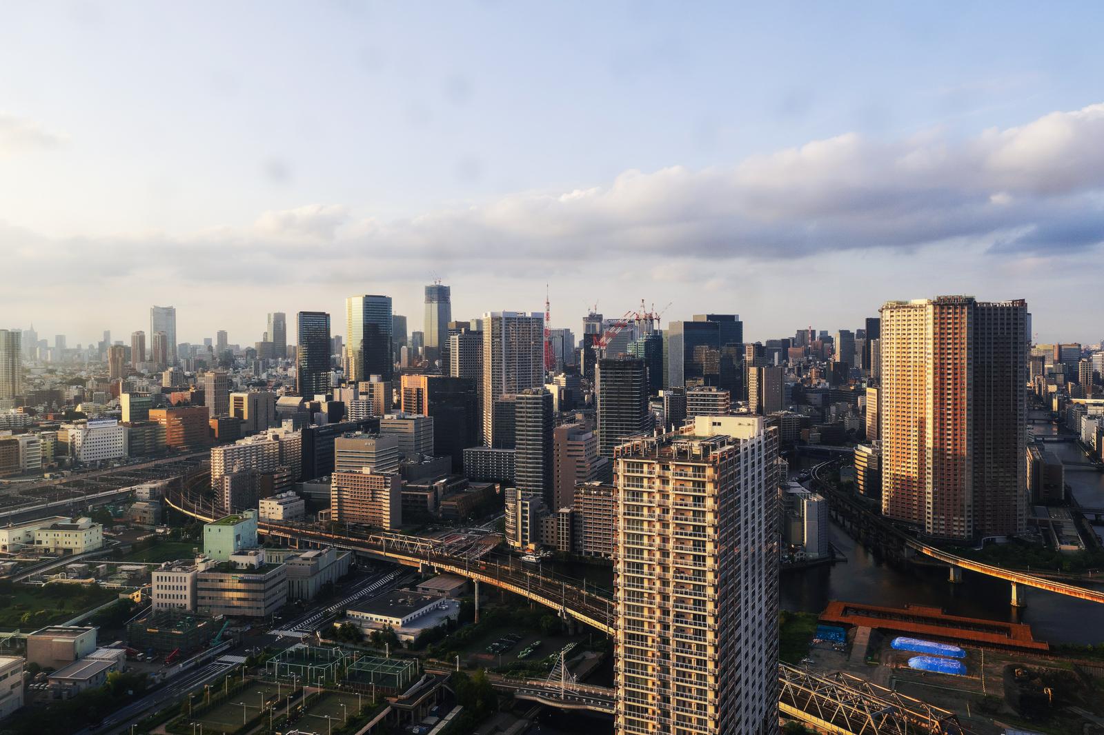 Aerial view of Tokyo skyline at golden hour showing high-rise towers, elevated highways, canals, and Tokyo Tower under a partly cloudy blue sky.