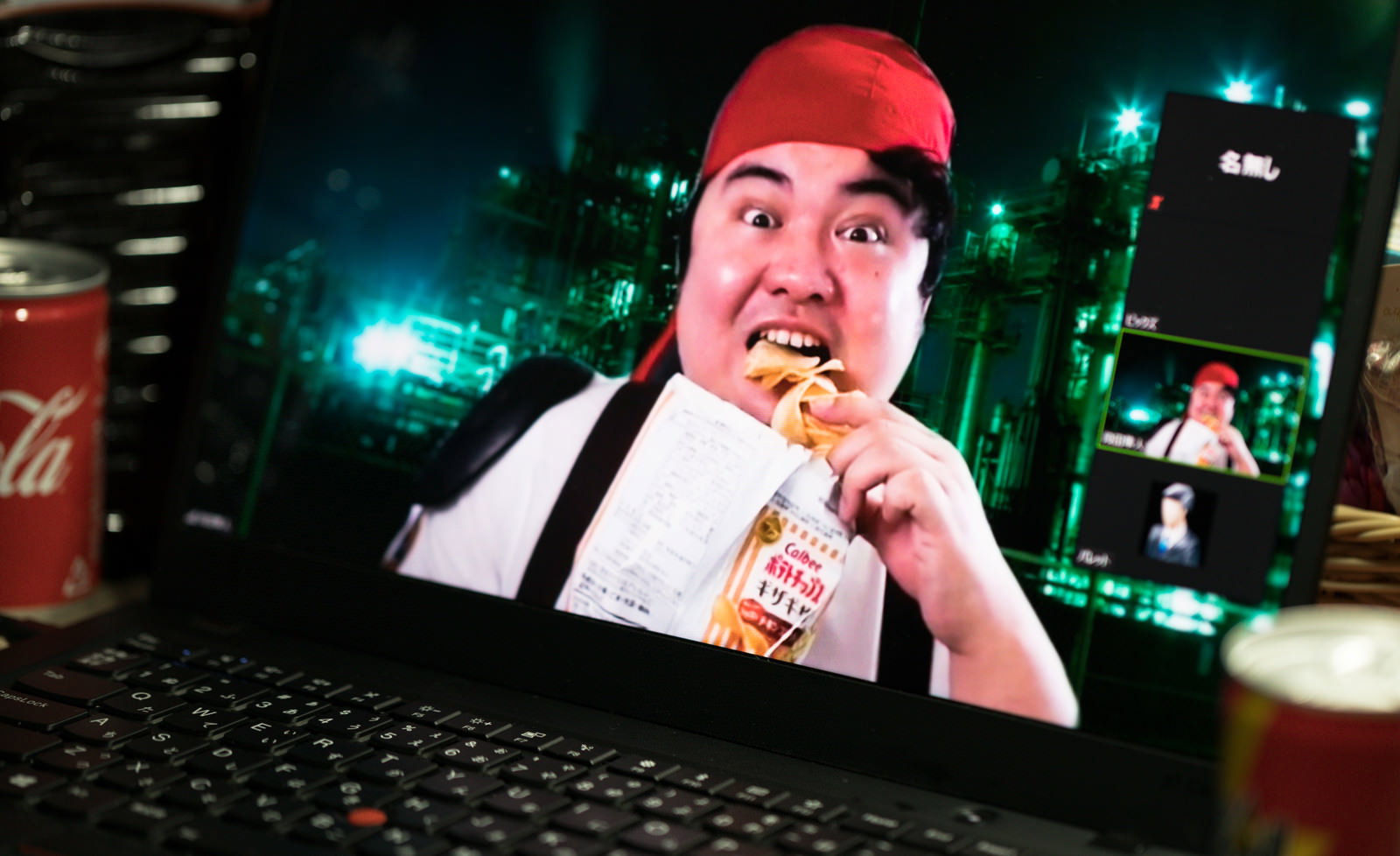 赤い帽子をかぶった男性がパソコンでビデオ通話をしながらポテトチップスを食べている［モデル：段田隼人］