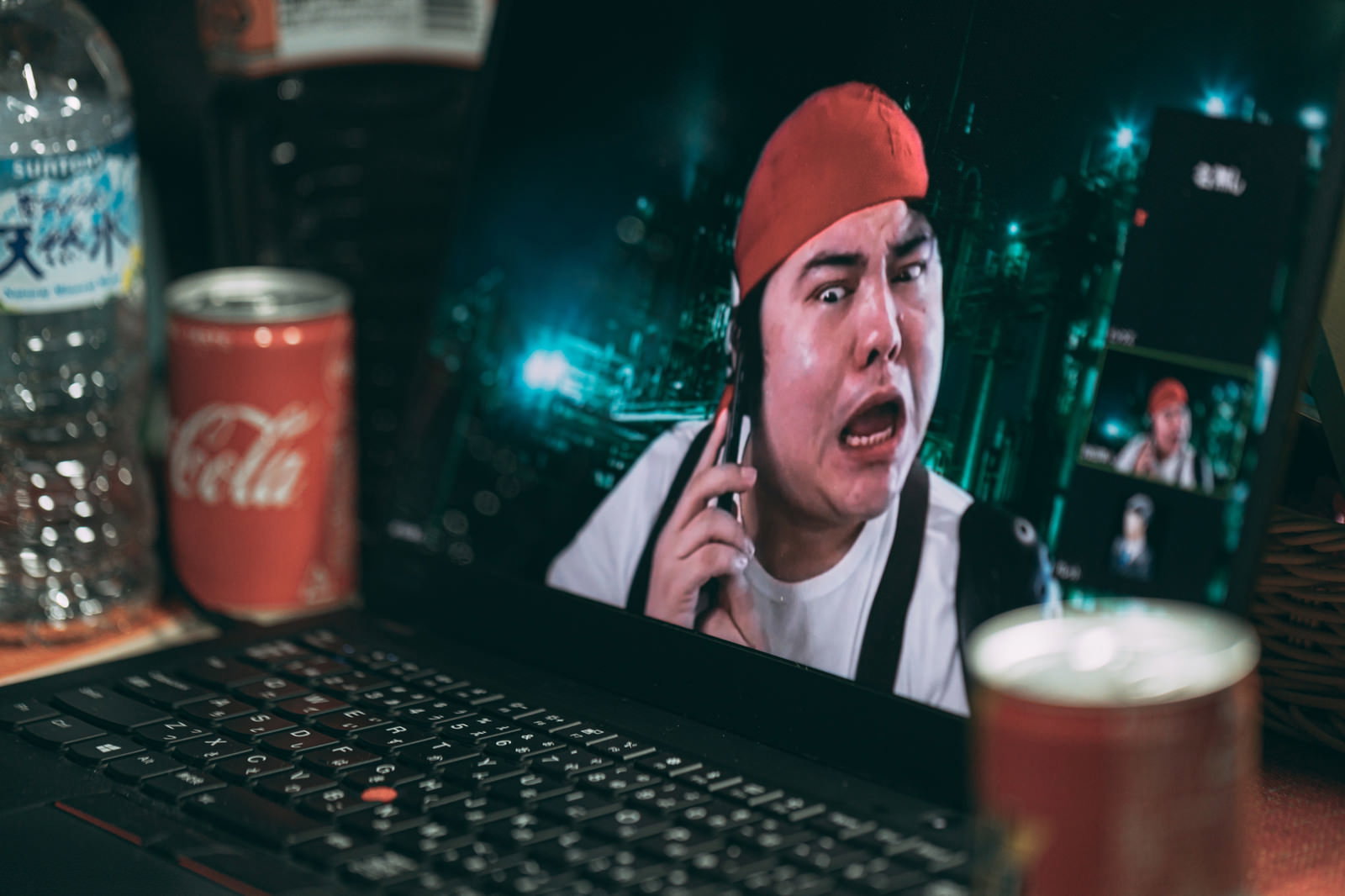 赤いバンダナの男性がパソコン前でスマートフォンを持ち困惑している様子［モデル：段田隼人］