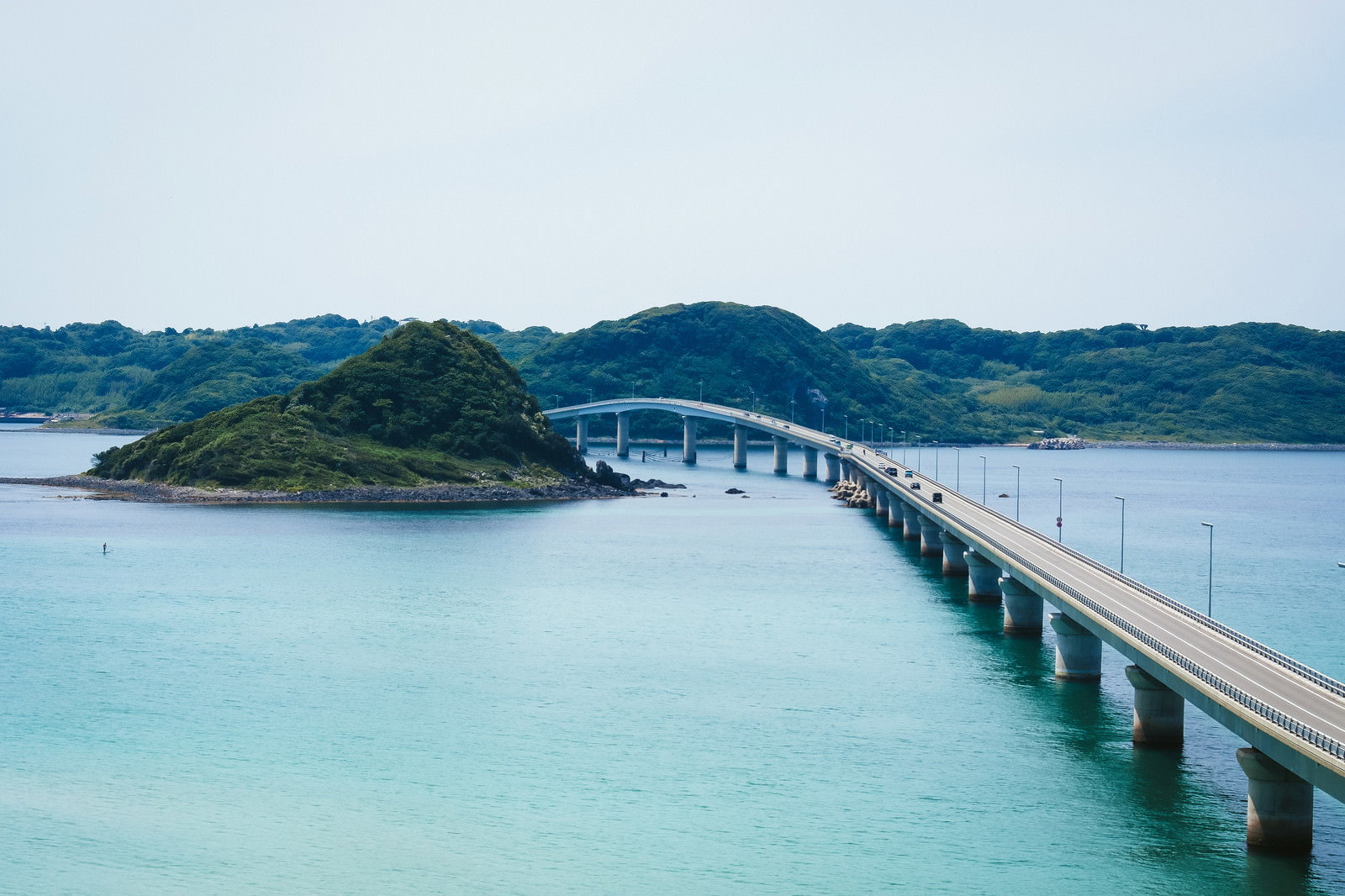 白いアーチ橋が緑の島を越えて海に架かり、背景に緑の山々がある景観