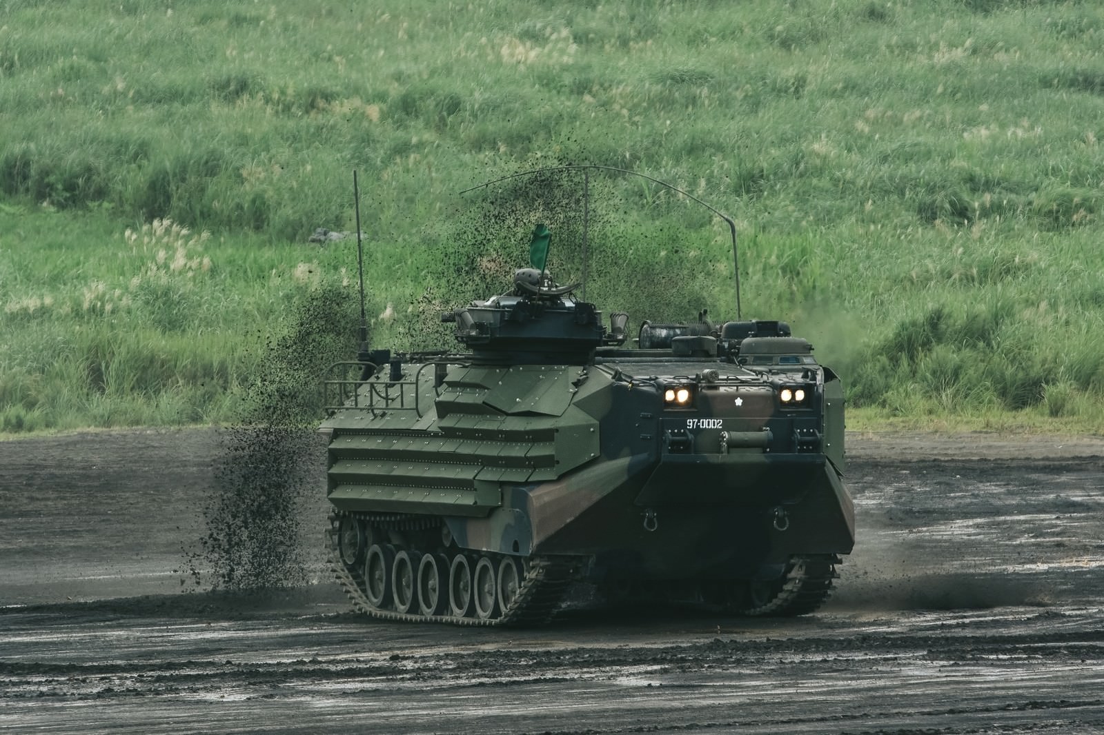 演習場の泥道を走行し泥を巻き上げる迷彩色のAAV7水陸両用装甲車両