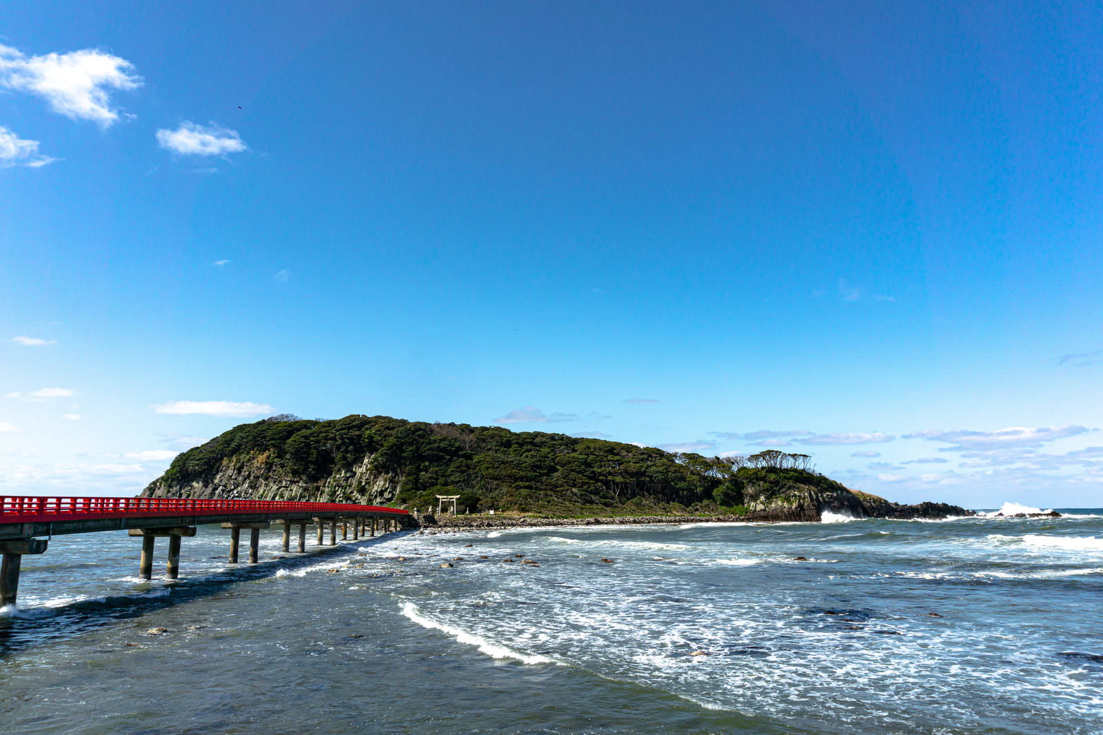 緑の島に赤い橋がかかり、白い波が押し寄せる海岸の風景