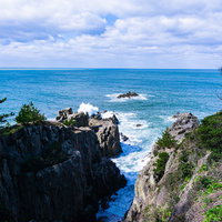 切り立った岩壁の間で白く泡立つ荒れた波と三段岩に高くあがる波しぶきとその先の軍艦岩の写真