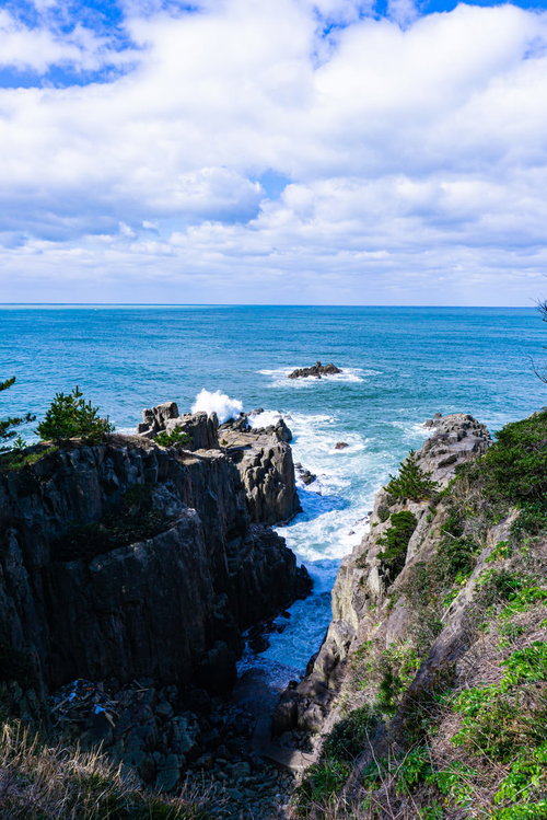 切り立った岩壁の間で白く泡立つ荒れた波と三段岩に高くあがる波しぶきとその先の軍艦岩