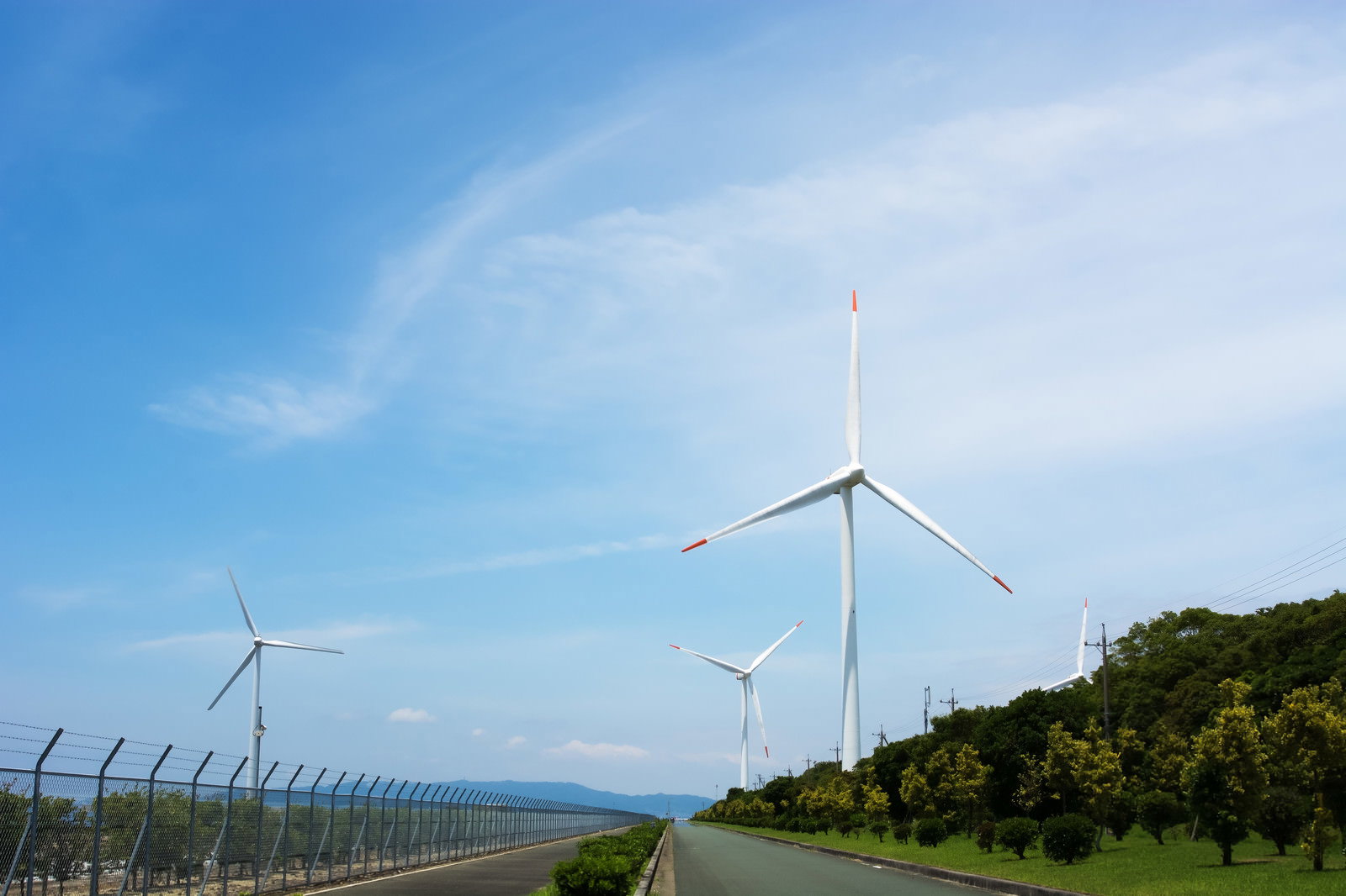 青空の下、緑の丘陵地帯に風力発電機が並び道路が続く風景