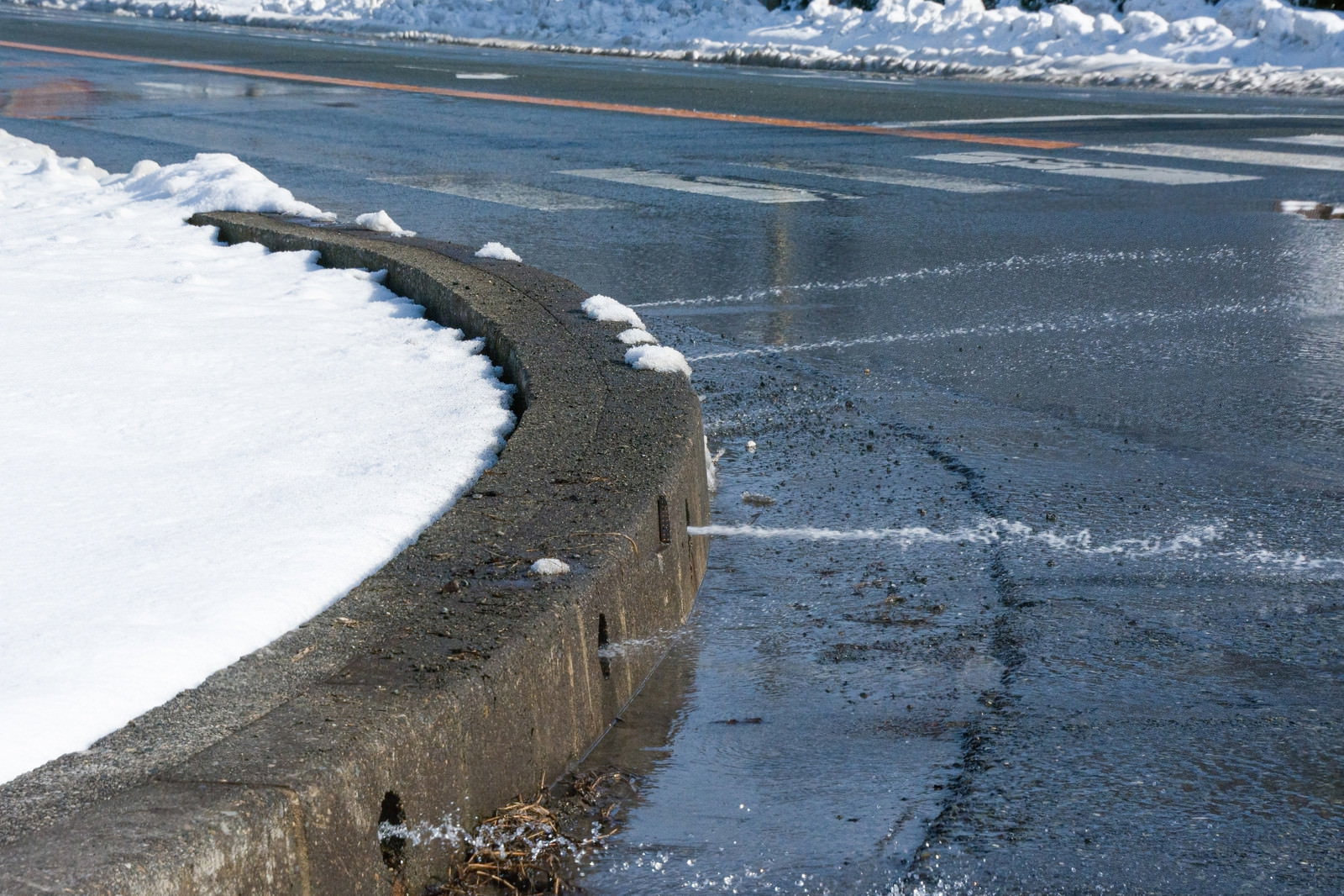 道路の縁石から水が流れ出し積もった雪を溶かしている様子