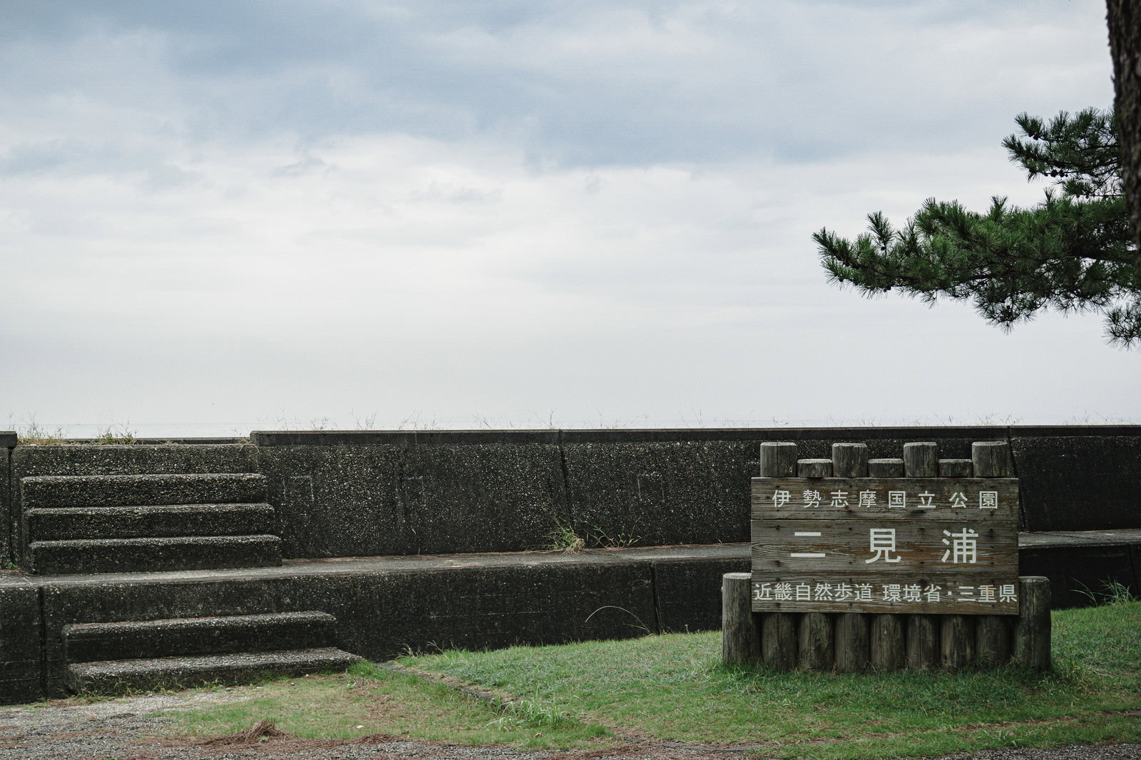 曇り空の下、石段と松の木がある屋外に設置された三重県二見浦の国立公園案内看板