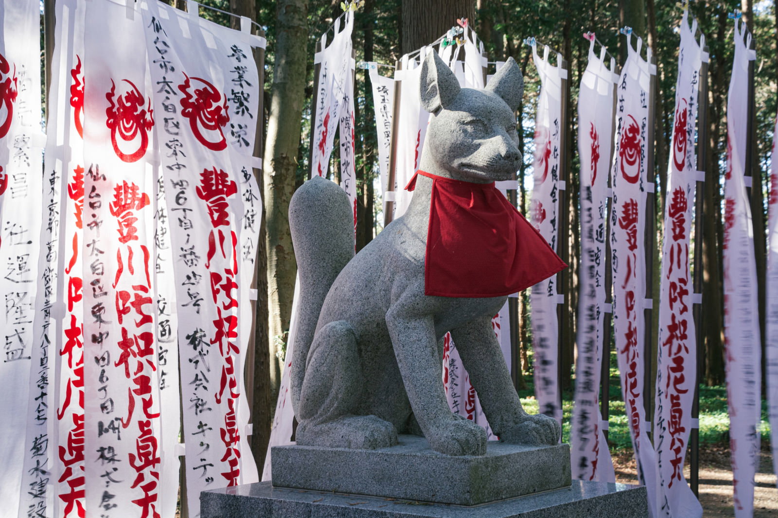 A white stone fox statue wearing a red bib, surrounded by upright white votive banners