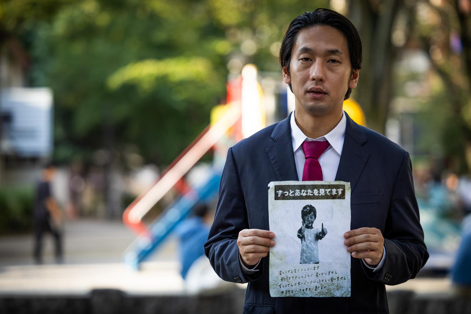 公園で「ずっとあなたを見てます」と書かれた怪文書を両手で掲げるスーツ姿の男性