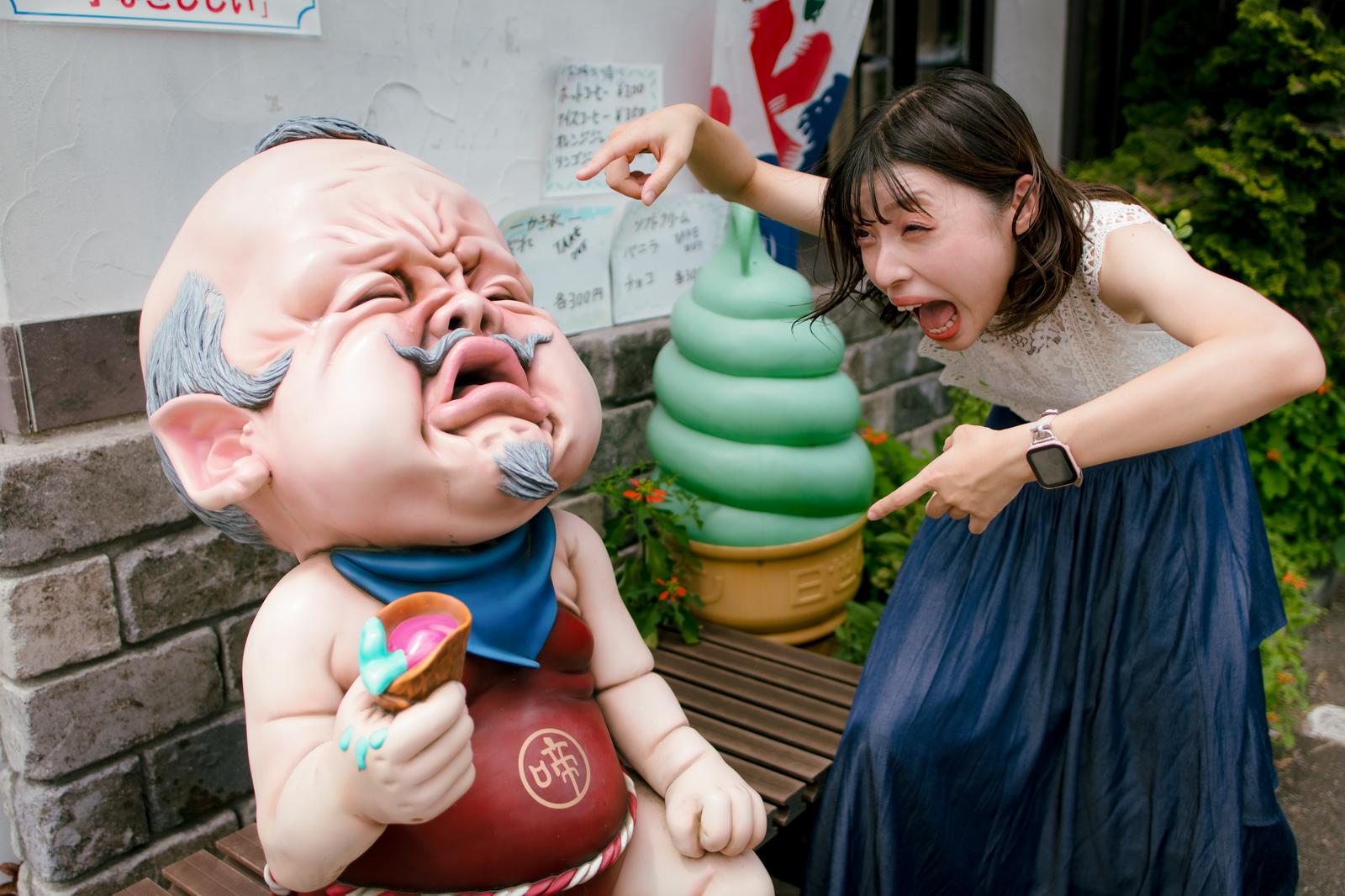 妖怪ベンチの子啼爺のオブジェを前にプギャーと煽る女性［モデル：高尾実生］