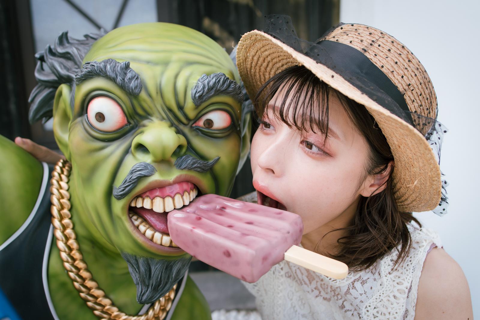 緑色の妖怪オブジェの横で麦わら帽子の女性がピンク色のアイスを食べている写真［モデル：高尾実生］
